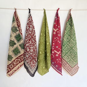 May include: Five colorful bandanas hang against a white background. The bandanas feature various patterns, including turtles, paisley, and geometric designs, in shades of beige, red, green, and purple. Each bandana is knotted at the top and suspended from a string.
