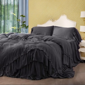 May include: A dark gray bed set with a ruffled bed skirt, duvet cover, and pillowcases. The bedding is on a bed with a white headboard and a gold-colored wall in the background. The room has a window with green curtains.