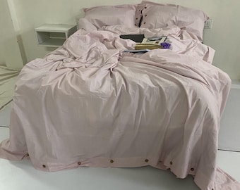 May include: A bed covered in a light pink duvet and pillow set. The bedding is slightly rumpled, with visible wooden buttons along the duvet's edge. A book and a small bouquet of purple flowers rest on the bed.