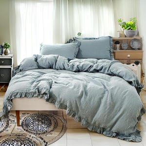 May include: A pale blue bed set with frilled edging. The set includes a duvet cover and pillowcases. The bed is made with a soft, textured fabric. The room has white curtains and a small bedside table.