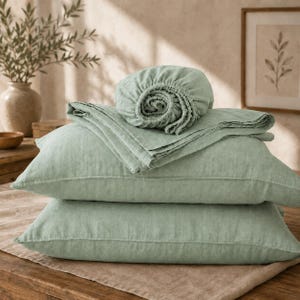 May include: A set of sage green bedding featuring two rectangular pillowcases stacked horizontally, topped with a folded flat sheet and a gathered fitted sheet. The fabric has a visible woven texture and a soft, matte finish in a muted green tone.