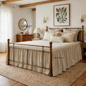 May include: A bedroom scene featuring a brass bed with beige bedding and a matching bed skirt. The room includes a jute rug, wooden furniture, and a framed botanical print. Natural light enters through a large window, creating a warm and inviting atmosphere.