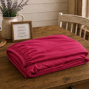 May include: A folded, deep pink fitted sheet rests on a wooden table. A small framed sign with the words "HOME SWEET HOME" is next to the sheet. A lantern and a vase with lavender are also on the table.