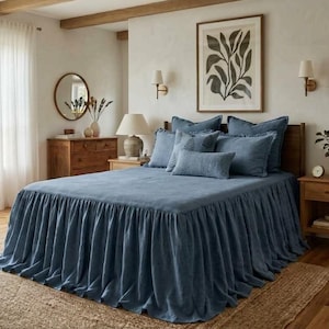 May include: A bedroom with a blue bedspread and matching pillows. The bed has a ruffled skirt and is set against a wooden headboard. A framed botanical print hangs above the bed. The room features wooden furniture, a round mirror, and a jute rug.