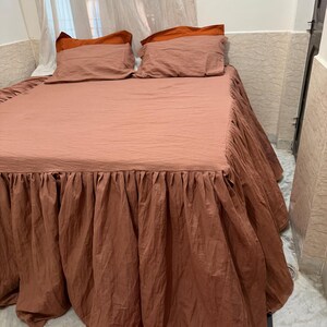 May include: A bed with a brown bedspread and matching ruffled bed skirt. Two pillows with rust-colored pillowcases are on the bed. The bed is in a room with a window and white curtains.