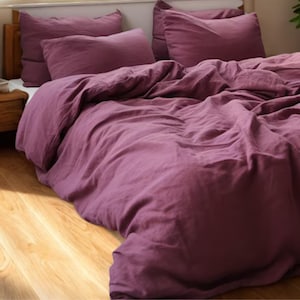 Może przedstawiać: Łóżko z ciemnofioletową lnianą poszwą na kołdrę i pasującymi poszewkami na poduszki. Pościel ułożona jest na drewnianej ramie łóżka, z drewnianą szafką nocną widoczną po lewej stronie. Podłoga jest z jasnego drewna.