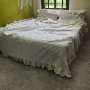 May include: A white bed with a ruffled bed skirt and matching ruffled pillowcases. The bed is covered with a white comforter. The room has a green wall and a window with a view of trees.