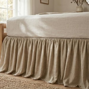 May include: A bed with a beige linen bed skirt and matching bedding. The bed skirt has a pleated design, and the bed frame is made of light-colored wood. A small vase sits on the floor near the bed.
