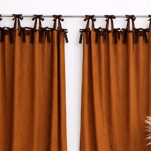 Puede incluir: Dos cortinas de lino color óxido cuelgan de una barra plateada. Cada cortina está adornada con lazos marrón oscuro en la parte superior. Las cortinas están ligeramente recogidas, creando pliegues suaves. La estética general es cálida y acogedora.