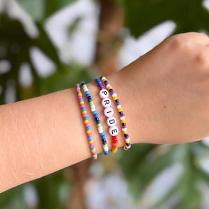 Könnte beinhalten: Vier Armbänder mit Perlen an einem Handgelenk. Die Armbänder sind mehrfarbig, eines buchstabiert "PRIDE" mit weißen Buchstabenperlen. Die anderen Armbänder haben Regenbogen-, blaue und violette Perlen. Die Armbänder befinden sich am Handgelenk einer Person.