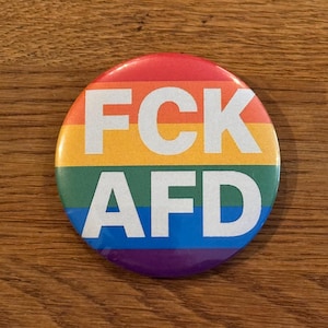 Könnte beinhalten: Ein runder Button mit Regenbogen-Farbverlauf und dem weißen Text "FCK AFD". Der Button liegt auf einer Holzoberfläche.
