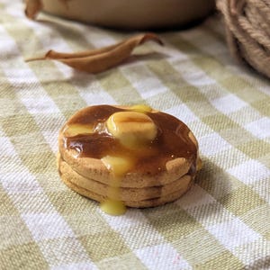 May include: A miniature stack of three pancakes, drizzled with syrup and topped with butter. The pancakes sit on a green and white checked cloth. A jar labelled "TEABAGS" is in the background.