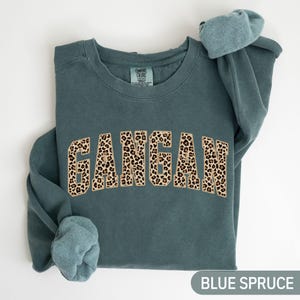 May include: Blue spruce sweatshirt with the word "GANGAN" in a leopard print design. The sweatshirt has a crew neck and long sleeves. The text "BLUE SPRUCE" is at the bottom of the image.