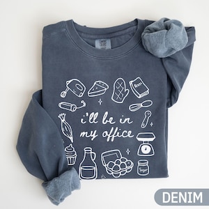 May include: Denim blue sweatshirt with white line art of baking tools and the phrase "I'll be in my office." Includes a mixer, pie, oven mitts, recipe book, whisk, scale, and more. The word "Denim" is printed at the bottom.