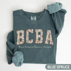 May include: Blue spruce sweatshirt featuring the letters "BCBA" in a floral pattern. The words "Board Certified Behavior Analyst" are printed below. The sweatshirt is made of a soft material.