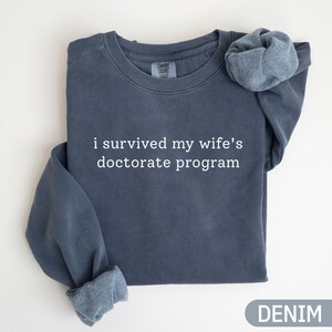 May include: A denim blue sweatshirt with the text "i survived my wife's doctorate program" in white. The sweatshirt has a crew neck and long sleeves with cuffed ends. The word "DENIM" is printed at the bottom.