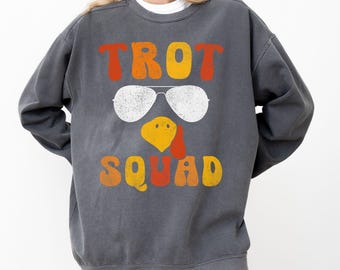 May include: A dark grey sweatshirt featuring the words "TROT SQUAD" in orange and yellow retro font. The design includes white sunglasses and a yellow and orange turkey head. The word "PEPPER" is in a white box at the bottom.