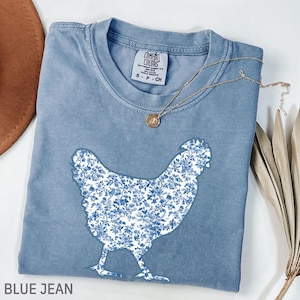 May include: A blue jean Comfort Colors t-shirt featuring a white chicken silhouette filled with a blue floral pattern. A gold necklace with a pendant rests on the shirt. The text "BLUE JEAN" is at the bottom.
