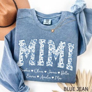 以下が含まれることがあります： 青いジーンズ色のスウェットシャツ。大きな白い文字で「MIMI」と書かれており、青い花柄で埋められています。名前の下には、Sophia、Oliver、James、Bella、Emma、Amelia、Maxの名前があります。金のネックレスがスウェットシャツにかけられています。