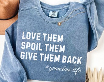 May include: Blue jean-colored sweatshirt with the text "LOVE THEM SPOIL THEM GIVE THEM BACK #grandma life" in white. A gold necklace with a pendant and a brown hat are also visible.