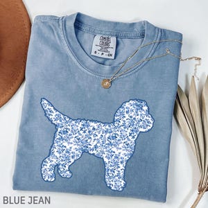 Peut inclure: T-shirt bleu jean avec une silhouette de chien à motifs floraux. Le t-shirt a un col rond et des manches courtes. Un collier en or avec un pendentif est drapé sur le t-shirt. Le texte "BLUE JEAN" est au bas de l'image.