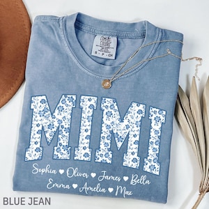 Könnte beinhalten: Ein blaues Comfort Colors T-Shirt mit dem Wort "MIMI" in großen, weißen Buchstaben, die mit einem blauen Blumenmuster gefüllt sind. Unter dem Wort "MIMI" stehen die Namen Sophia, Oliver, James, Bella, Emma, Amelia und Max. Eine goldene Halskette liegt auf dem Shirt.