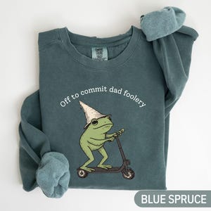 Peut inclure: Un sweat-shirt bleu épicéa à col rond orné d'un motif représentant une grenouille verte portant un chapeau pointu beige et montant une trottinette noire. Le texte au-dessus de la grenouille indique Off to commit dad foolery en blanc.