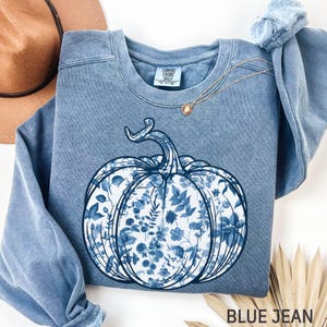 May include: A blue jean-coloured sweatshirt with a large pumpkin design filled with blue floral patterns. The sweatshirt has long sleeves and a crew neck. The text "BLUE JEAN" is at the bottom of the image.