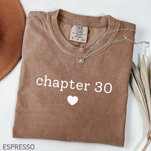 May include: An espresso-colored t-shirt with a crew neckline and short sleeves. The front features the white text 'chapter 30' above a small white heart graphic. A silver-toned necklace with a circular pendant rests on the fabric.