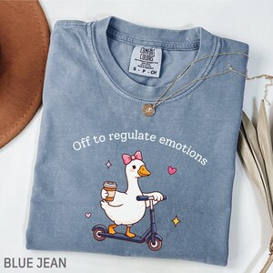 May include: A blue jean coloured t-shirt featuring the text "Off to regulate emotions" and a cartoon goose on a scooter with a coffee cup. A gold necklace and a small tag are visible. The shirt is folded.