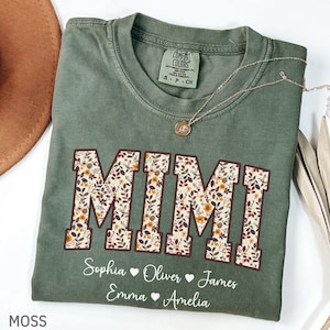 以下が含まれることがあります： モスグリーンのTシャツ。大きな花柄の文字で「MIMI」と書かれています。その下に、Sophia、Oliver、James、Emma、Ameliaという名前があります。ペンダント付きのシルバーネックレスがシャツの上にあります。