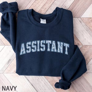 Op de afbeelding: Marineblauwe sweatshirt met het woord "ASSISTANT" in een wit en blauw bloemmotief op de borst. Het woord "NAVY" staat onderaan afgedrukt. De sweatshirt heeft een ronde hals en lange mouwen.