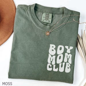 以下が含まれることがあります： モスグリーンのTシャツ。白のレトロな文字で「BOY MOM CLUB」と書かれています。ペンダント付きのゴールドのネックレスがシャツの上にかけられています。シャツは折りたたまれており、背景には茶色の帽子が見えます。