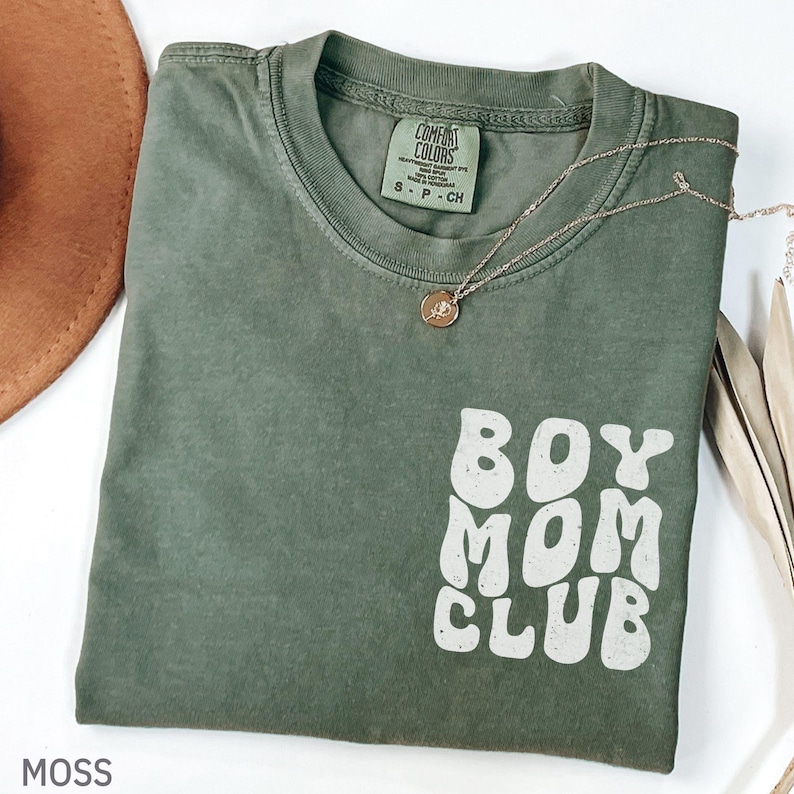 以下が含まれることがあります： モスグリーンのTシャツ。白のレトロな文字で「BOY MOM CLUB」と書かれています。ペンダント付きのゴールドのネックレスがシャツの上にかけられています。シャツは折りたたまれており、背景には茶色の帽子が見えます。
