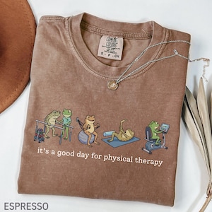 May include: An espresso-colored t-shirt with a graphic print of five green frogs performing physical therapy exercises. The text below the frogs reads, it's a good day for physical therapy. The shirt features a Comfort Colors brand neck label.