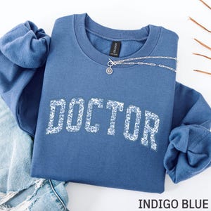 May include: Indigo blue sweatshirt with the word "DOCTOR" in white floral print. A silver necklace with a sunburst pendant is draped across the top. The sweatshirt is folded on top of a pair of blue jeans.