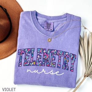 May include: A violet Comfort Colors t-shirt with the word "TELEMETRY" in floral print and the word "nurse" in white script. A gold necklace with a pendant rests on the shirt. A brown hat and dried plant are in the background.