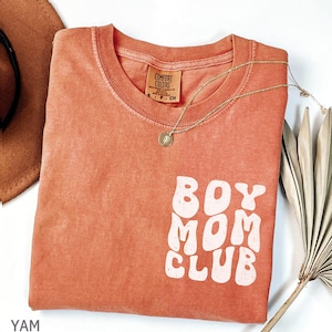 以下が含まれることがあります： 白いブロック文字で「BOY MOM CLUB」と書かれたコーラルカラーのTシャツ。 金色のネックレスとペンダントがシャツの上にあります。 シャツは折りたたまれており、「COMFORT COLORS」と書かれたタグが付いています。
