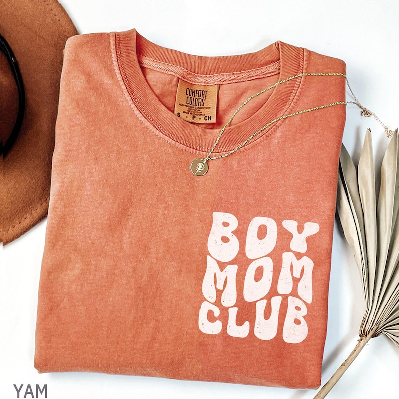 以下が含まれることがあります： 白いブロック文字で「BOY MOM CLUB」と書かれたコーラルカラーのTシャツ。 金色のネックレスとペンダントがシャツの上にあります。 シャツは折りたたまれており、「COMFORT COLORS」と書かれたタグが付いています。