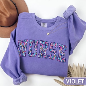 May include: A violet sweatshirt featuring the word "NURSE" in a floral design across the chest. The long-sleeved top has a crew neck and a gold necklace. The word "VIOLET" is printed in a white rectangle at the bottom right.