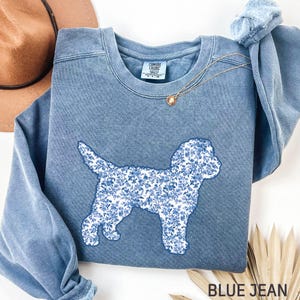 May include: Blue jean-colored sweatshirt with a floral dog silhouette. The dog design is filled with a blue and white floral pattern. A gold necklace with a pendant is draped across the sweatshirt. The text "BLUE JEAN" is at the bottom.