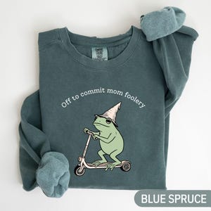 Peut inclure: Un sweat-shirt bleu-vert à col rond avec une illustration d'une grenouille vert clair portant un chapeau pointu et montant une trottinette blanche. Le texte au-dessus de la grenouille indique, Off to commit mom foolery. Manches longues.