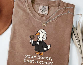 May include: Espresso-colored Comfort Colors t-shirt featuring a cartoon duck dressed as a judge, holding a gavel. The text "your honor, that's crazy" is printed below the duck. A silver necklace with a small pendant is draped across the shirt.
