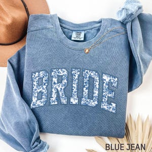 May include: A blue jean-coloured sweatshirt featuring the word "BRIDE" in large, white, floral-patterned letters. A gold necklace with a small pendant is draped on the sweatshirt. A brown hat is visible in the background.