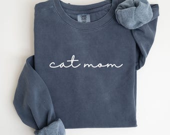 May include: A denim blue sweatshirt with the words "cat mom" in white script. The sweatshirt has a crew neck and long sleeves with folded cuffs. The word "DENIM" is printed in a white rectangle in the bottom right corner.