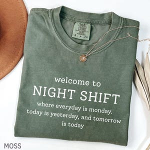 Puede incluir: Una camiseta verde musgo con el texto "welcome to NIGHT SHIFT" en blanco. La camiseta también tiene las frases "where everyday is monday. today is yesterday, and tomorrow is today." Un collar dorado con un colgante está en la camiseta.
