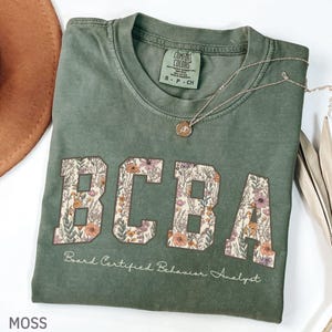 May include: A moss green t-shirt featuring the letters "BCBA" in a floral design. The text "Board Certified Behavior Analyst" is below. A silver necklace with a pendant rests on the folded shirt.