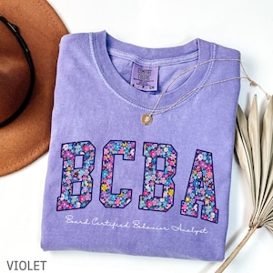 May include: A violet Comfort Colors t-shirt with the letters "BCBA" in a floral pattern. The text "Board Certified Behavior Analyst" is below the letters. A gold necklace and a brown hat are also in the image.
