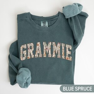 May include: A blue spruce sweatshirt featuring the word "GRAMMIE" in a floral pattern. The sleeves are rolled up. The text "BLUE SPRUCE" is at the bottom. The sweatshirt is folded.