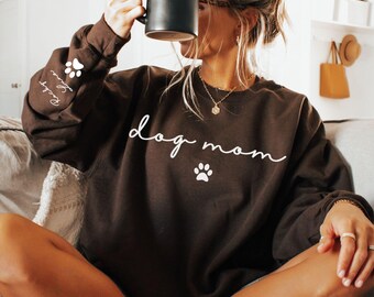 May include: Dark chocolate brown sweatshirt with the words "dog mom" in white script and a paw print graphic. The sleeve has paw prints and names. The bottom right corner of the image has the words "DARK CHOCOLATE".